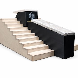 May include: A miniature skateboard ramp with a staircase and a ledge. The ramp features a light wood staircase, a black wall, and a concrete ledge. A silver emblem is on the black wall. The ramp is designed for fingerboarding.