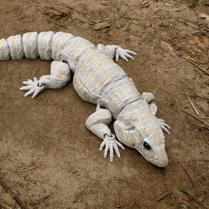May include: A detailed, off-white lizard sculpture with a segmented body and articulated limbs. The reptile has a textured surface with subtle yellow highlights and green eyes. It is lying on a bed of brown earth and small twigs.