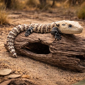 Peut inclure: Un monstre de Gila se repose sur une bûche patinée. Le lézard a un corps crème avec des marques marron et noires. Ses griffes noires agrippent le bois, et il a un petit œil perlé. La bûche est d'une riche couleur marron, et le fond est de couleur sable.