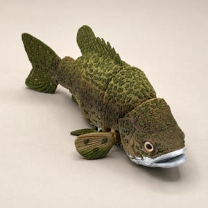 May include: A realistic fish figurine featuring a textured olive green and brown mottled body with a prominent dorsal fin, a light-colored underbelly, and a silver-toned mouth. The fish is depicted in a swimming position with detailed scales.