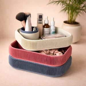 Puede incluir: Una pila de tres bandejas de almacenamiento acanaladas en crema, rojo y azul marino. La bandeja superior contiene brochas de maquillaje, productos para el cuidado de la piel y un pequeño recipiente azul. Una planta en maceta está en el fondo.