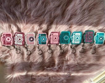 May include: A collection of camera-shaped keychains in various colors, including pink, teal, and white, with glitter accents. Each keychain has a silver ring. The keychains are arranged on a pink, fluffy surface.