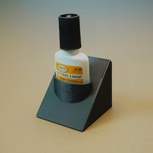 May include: A black 3D printed holder for a bottle of fly tying cement. The bottle is white with a black cap and yellow label that reads "Wapsi Fly Tyers Z-Ment Wapsi Formula PF-80 A49A".