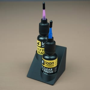 May include: Two bottles of Loon Outdoors fly tying finish, one black and one clear, in a black plastic holder. The bottles have a black cap with a colored applicator tip, one purple and one blue.