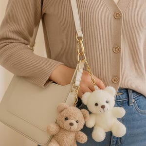May include: Two plush teddy bear keychains, one beige and one white, hang from a cream-colored purse. The bears have dark brown eyes and noses. The purse has a gold chain and a white strap. The background includes a beige cardigan and blue jeans.