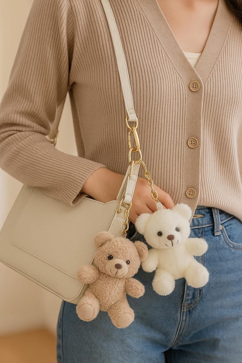 May include: Two plush teddy bear keychains, one beige and one white, hang from a cream-colored purse. The bears have dark brown eyes and noses. The purse has a gold chain and a white strap. The background includes a beige cardigan and blue jeans.