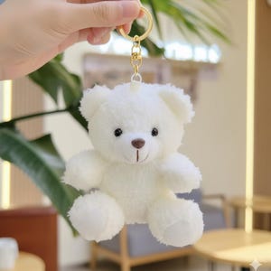May include: A small, fluffy white teddy bear keychain with a gold-colored keyring. The bear has black eyes, a brown nose, and a stitched smile. The plush toy is soft and cuddly, perfect for attaching to keys or a backpack.