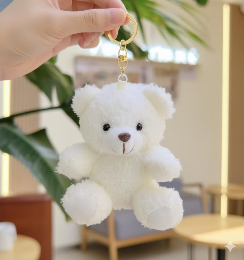 May include: A small, fluffy white teddy bear keychain with a gold-colored keyring. The bear has black eyes, a brown nose, and a stitched smile. The plush toy is soft and cuddly, perfect for attaching to keys or a backpack.