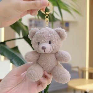 May include: A small, plush teddy bear keychain in a light brown color. The bear has a stitched nose and mouth, with small black eyes. The keychain has a gold-colored metal ring and clasp.