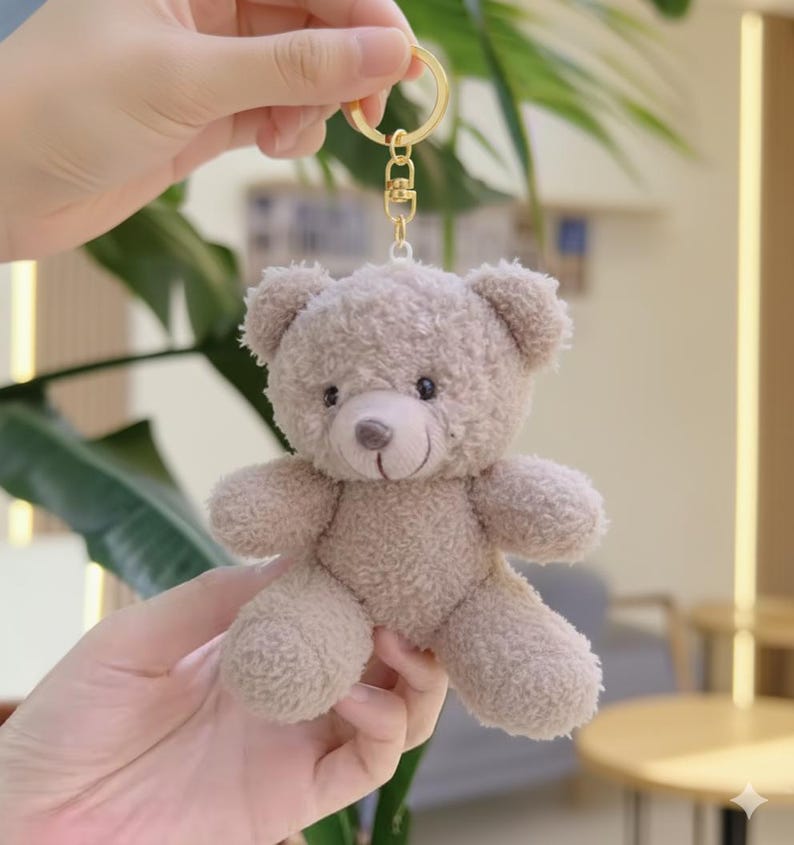 May include: A small, plush teddy bear keychain in a light brown color. The bear has a stitched nose and mouth, with small black eyes. The keychain has a gold-colored metal ring and clasp.