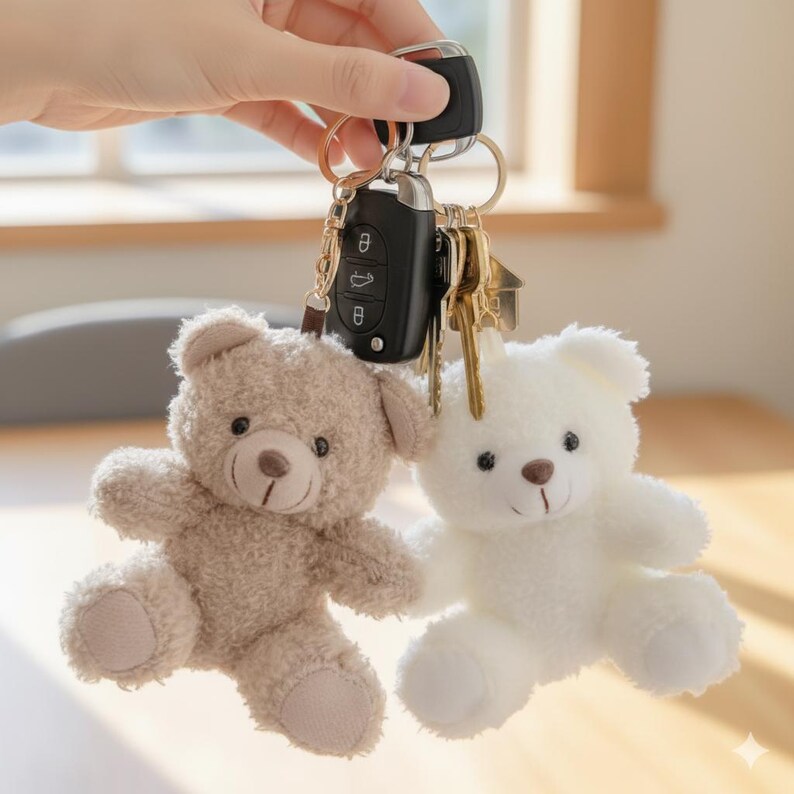 May include: Two plush teddy bear keychains, one tan and one white, hanging with keys and a car remote. The bears have brown eyes and noses. The keychains are held by a hand.