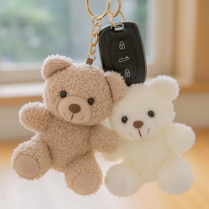 May include: Two plush teddy bear keychains in beige and white, attached to a gold-colored keyring and a car key. The bears have black eyes and brown noses. The beige bear has a textured appearance.
