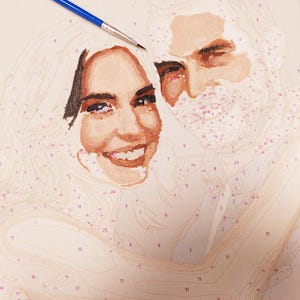 May include: A paint-by-numbers portrait in progress, featuring a smiling couple. The woman has blonde hair and the man has a beard. A blue paintbrush is positioned near the top of the canvas, with numbers and outlines visible.