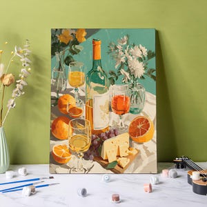 Puede incluir: Una pintura de naturaleza muerta representa una botella de vino, copas de vino, naranjas, uvas y queso. La pintura tiene un fondo verde claro con flores blancas y amarillas en jarrones. Pinceles y botes de pintura en primer plano.