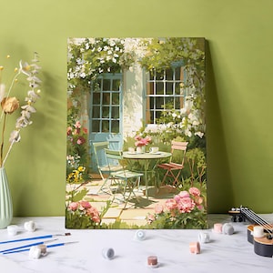Puede incluir: Una pintura enmarcada representa una encantadora escena de café al aire libre. Una mesa redonda y sillas están dispuestas en un patio, rodeadas de flores y vegetación. La pintura presenta una puerta y una ventana azules, con una paleta de colores suave y acogedora.