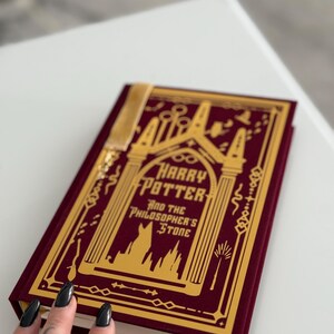 Könnte beinhalten: Ein Hardcover-Buch in burgunderfarbenem Samt mit einem goldenen Lesezeichen. Der Buchumschlag zeigt eine goldene Illustration von Hogwarts und den Text "Harry Potter und der Stein der Weisen".