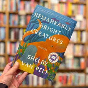 Könnte beinhalten: Buchcover von "Remarkably Bright Creatures" von Shelby Van Pelt. Es zeigt eine orangefarbene Oktopus-Illustration auf blauem Hintergrund. Der Titel ist weiß und der Name des Autors gelb. Ein "Read with Jenna"-Aufkleber ist ebenfalls sichtbar.