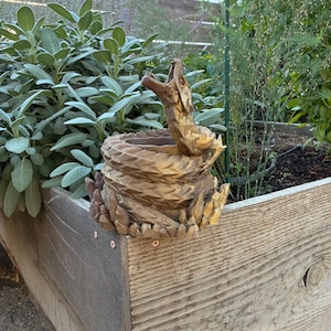 Op de afbeelding: Een gedetailleerde, goudkleurige slangensculptuur met een open bek, opgerold rond een cirkelvormige basis. De sculptuur staat op een houten plantenbak, omringd door groene planten. De textuur en de open bek van de slang zijn prominent aanwezig.