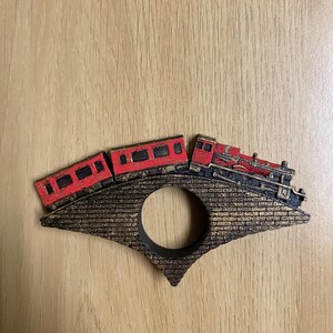 May include: A wooden train engine and two train cars painted red and black, crossing a brick bridge with a gold finish. The bridge has a circular opening in the center.