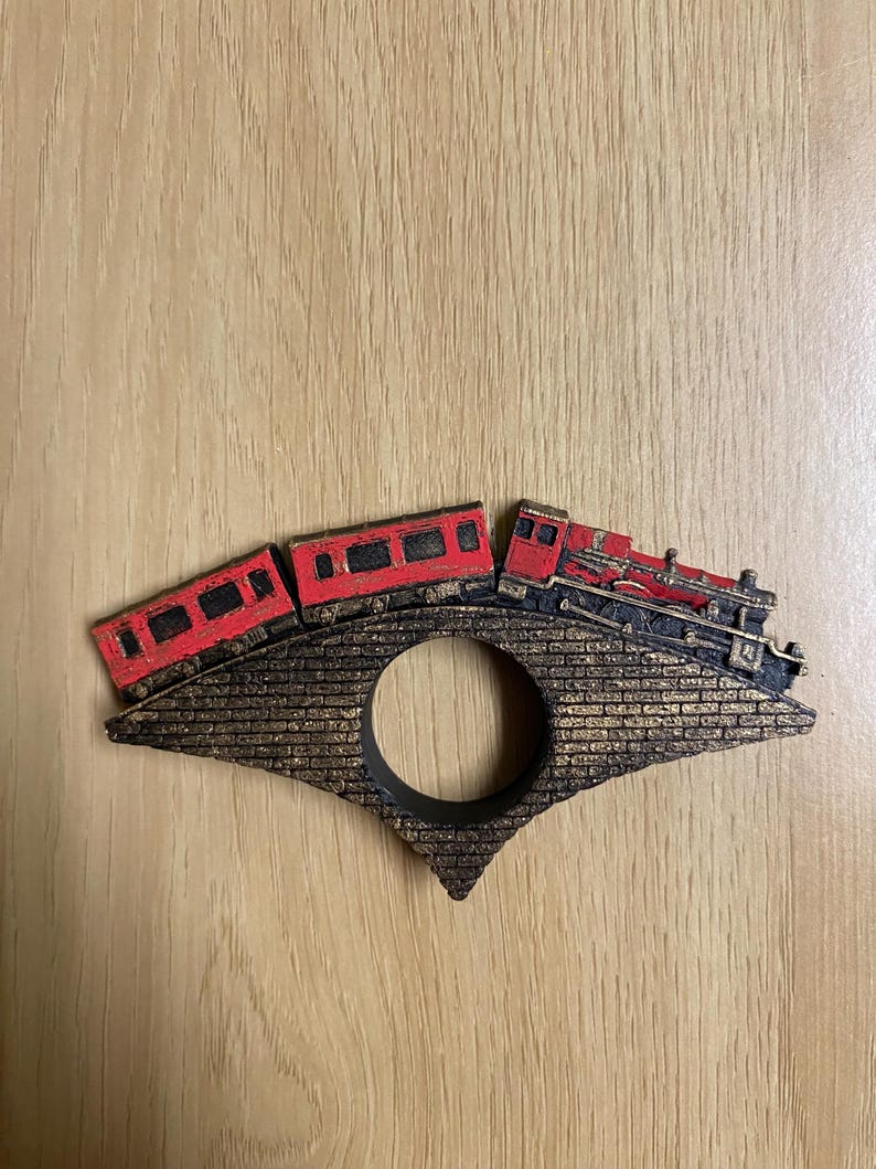 May include: A wooden train engine and two train cars painted red and black, crossing a brick bridge with a gold finish. The bridge has a circular opening in the center.