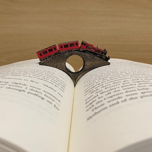Puede incluir: Una locomotora roja y dos vagones de tren cruzan un puente de madera. El puente es un marcapáginas que mantiene un libro abierto.