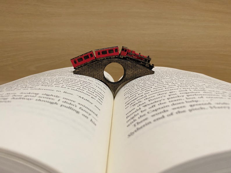May include: A red train engine and two train cars are crossing a bridge made of wood. The bridge is a bookmark that is holding open a book.