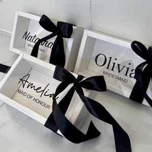 May include: Three white gift boxes with clear lids and black satin ribbon bows. The boxes are labelled with the names "Natasha, Bridesmaid", "Olivia, Bridesmaid", and "Amelia, Maid of Honour".