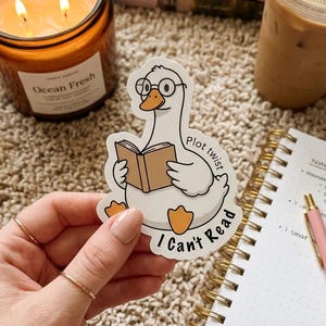 May include: A die-cut vinyl sticker featuring a white cartoon goose wearing round glasses and holding an open brown book. The text "Plot twist" and "I Can't Read" is printed in black around the goose. The sticker has a white border.