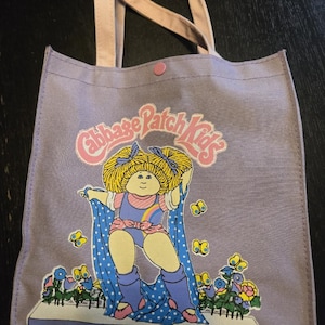 Peut inclure: Un sac fourre-tout lavande avec un motif Cabbage Patch Kids. Le dessin présente un personnage de dessin animé aux cheveux jaunes, un arc-en-ciel et des papillons. Le sac a des poignées roses et un bouton-pression rose.