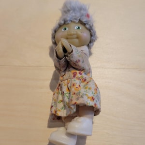 Puede incluir: Una muñeca pequeña con un gorro gris peludo y un vestido floral. La muñeca tiene ojos verdes y una cara sonriente. La muñeca tiene las manos juntas en posición de oración.