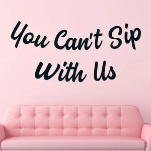May include: A pink sofa with button detailing is positioned beneath the black text "You Can't Sip With Us" on a pink wall. The room has a minimalist design with a white orb lamp. The overall theme is a modern, stylish living space.