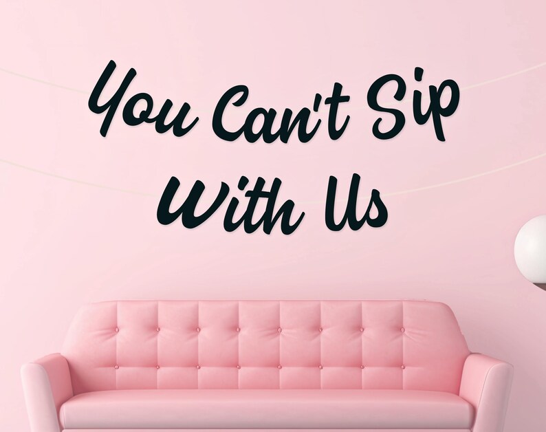 May include: A pink sofa with button detailing is positioned beneath the black text "You Can't Sip With Us" on a pink wall. The room has a minimalist design with a white orb lamp. The overall theme is a modern, stylish living space.