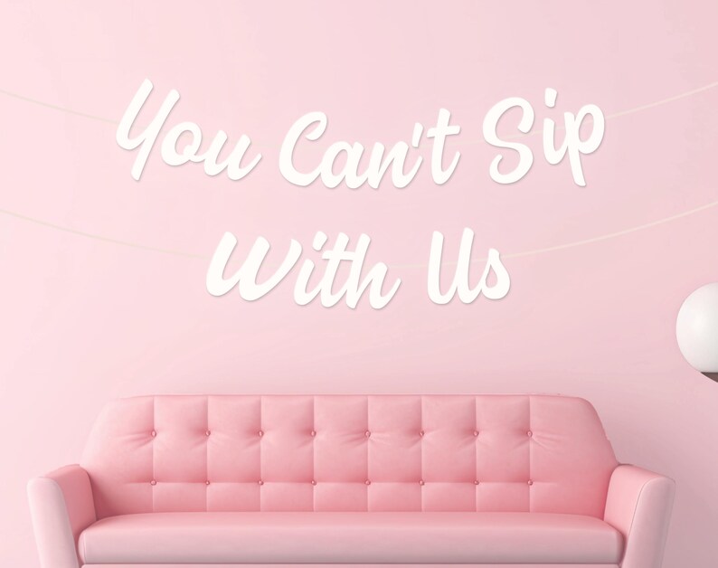 May include: A pink button-backed sofa is positioned beneath a banner with the white script text "You Can't Sip With Us". The background is a solid pink, with a white globe light on the right.