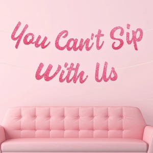 May include: Pink glitter banner reading "You Can't Sip With Us" suspended above a pink tufted sofa. The banner is strung across a light pink wall. A white orb lamp is visible on the right side of the image.