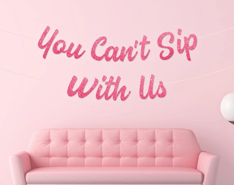 May include: Pink glitter banner reading "You Can't Sip With Us" suspended above a pink tufted sofa. The banner is strung across a light pink wall. A white orb lamp is visible on the right side of the image.