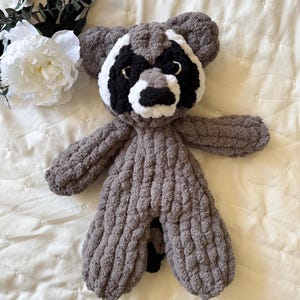 May include: A gray knitted raccoon plush toy with black eyes and a white nose. The toy is lying on a white quilted blanket.