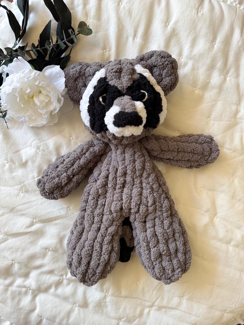 May include: A gray knitted raccoon plush toy with black eyes and a white nose. The toy is lying on a white quilted blanket.