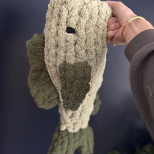 Puede incluir: Un juguete de peluche con forma de pez con cabeza y cola verdes y cuerpo beige. El pez tiene un ojo negro y est&aacute; hecho de una tela suave y texturizada.