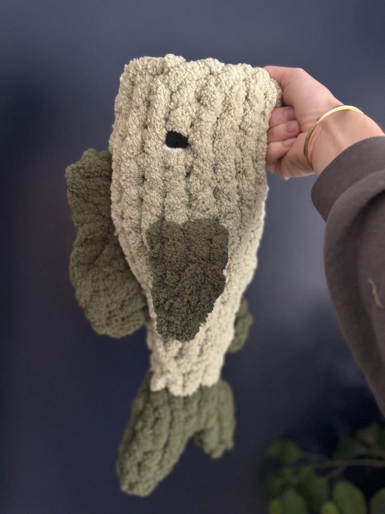 Puede incluir: Un juguete de peluche con forma de pez con cabeza y cola verdes y cuerpo beige. El pez tiene un ojo negro y est&aacute; hecho de una tela suave y texturizada.
