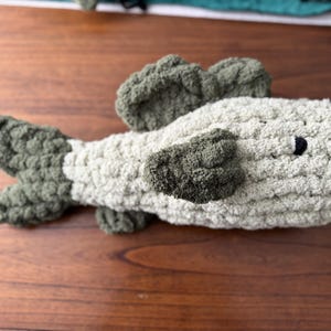 Puede incluir: Un juguete de peluche de crochet con forma de pez. El pez es de color verde claro y gris con un ojo negro.