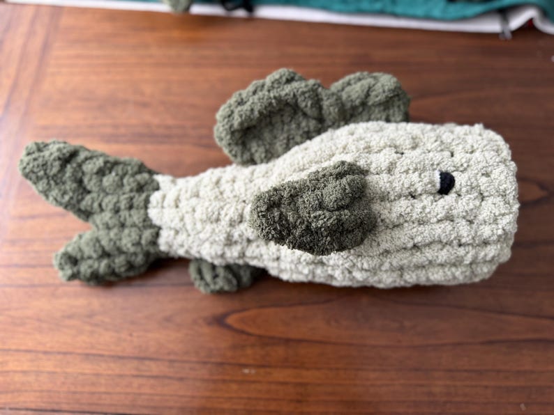 Puede incluir: Un juguete de peluche de crochet con forma de pez. El pez es de color verde claro y gris con un ojo negro.