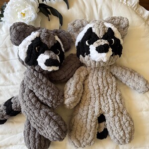 May include: Two plush raccoon toys, one gray and one tan, with black eyes and noses, and white markings around their eyes. The toys are sitting on a white, ruffled surface.