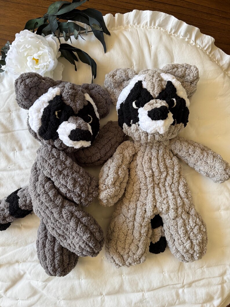 May include: Two plush raccoon toys, one gray and one tan, with black eyes and noses, and white markings around their eyes. The toys are sitting on a white, ruffled surface.