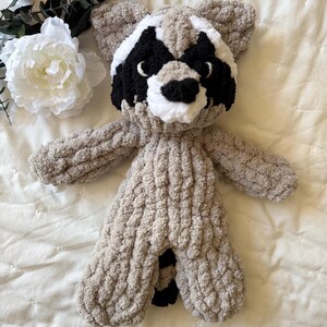 May include: A crocheted raccoon plush toy with a gray body, black eyes, and a white nose and mouth. The toy is lying on a white bedspread.