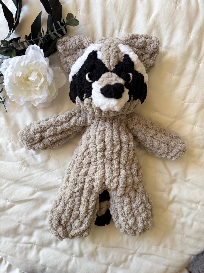 May include: A crocheted raccoon plush toy with a gray body, black eyes, and a white nose and mouth. The toy is lying on a white bedspread.