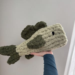 Puede incluir: Un juguete de peluche de ganchillo con forma de pez. El pez es verde y beige con un ojo negro.