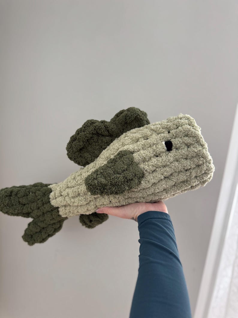 Puede incluir: Un juguete de peluche de ganchillo con forma de pez. El pez es verde y beige con un ojo negro.