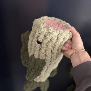 Puede incluir: Un juguete de peluche de crochet con forma de tronco de &aacute;rbol con una flor rosa en la parte superior. El juguete est&aacute; hecho de hilo suave y texturizado y tiene un esquema de color verde y marr&oacute;n.