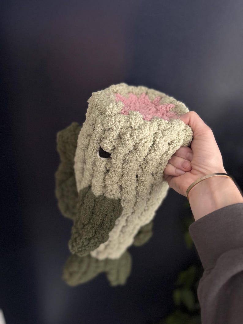 Puede incluir: Un juguete de peluche de crochet con forma de tronco de &aacute;rbol con una flor rosa en la parte superior. El juguete est&aacute; hecho de hilo suave y texturizado y tiene un esquema de color verde y marr&oacute;n.