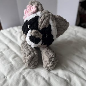 May include: A crocheted raccoon plush toy with a pink flower on its head. The toy is gray, black, and white and has a sweet expression.